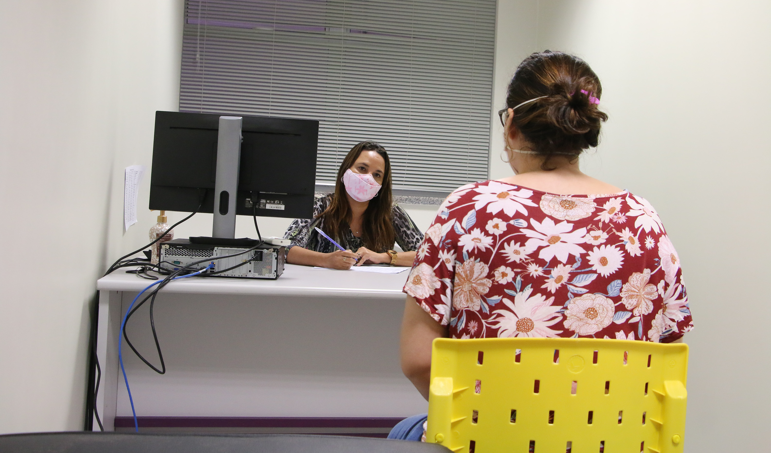 duas mulheres sentadas numa mesa conversando, ambas estão de máscara e uma delas está de costas para a câmera